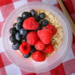 overnight oats with water and berries in clear container on checked cloth.