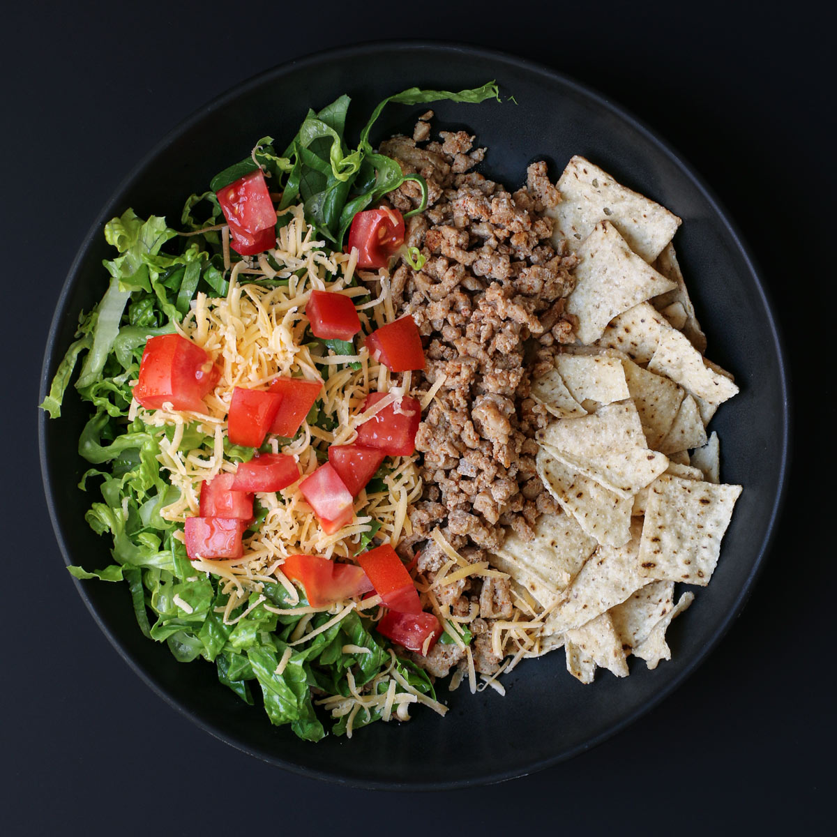 salad assembled in black bowl with chips, turkey meat, lettuce, cheese, and tomatoes.