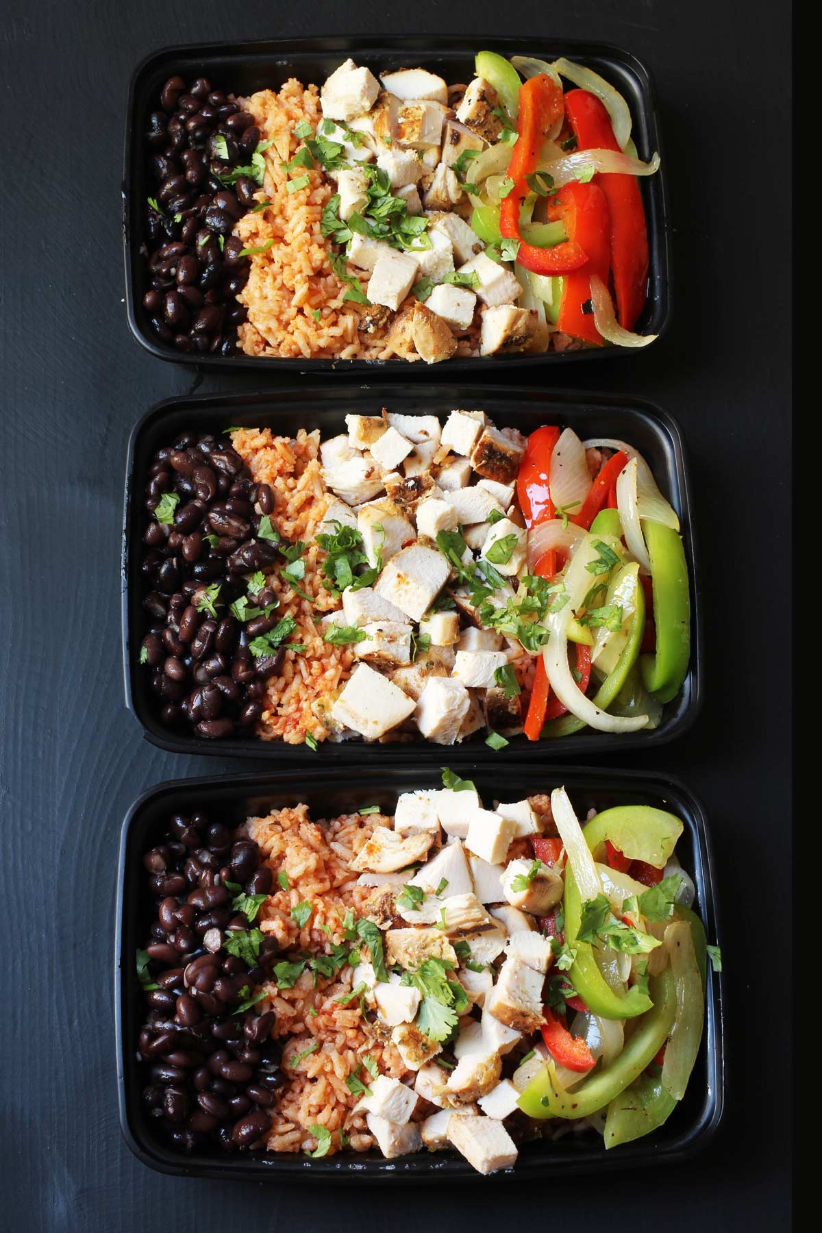 three black meal prep boxes with burrito rice bowls assembled in them, on a black table.