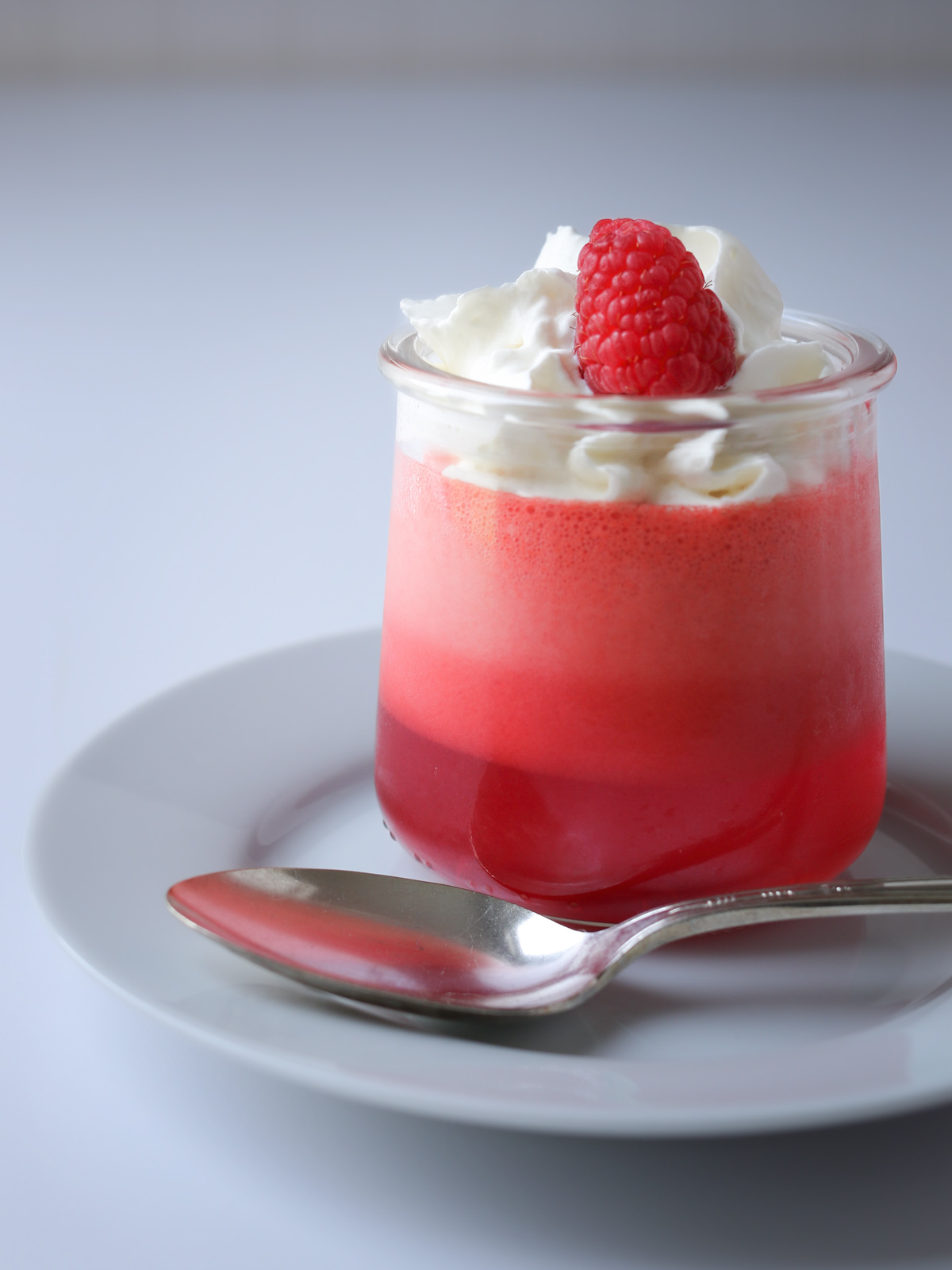 layered raspberry jello in glass ramekin on white plate, with spoon.