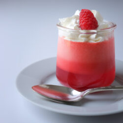 jello 123 copycat layered dessert in glass ramekin topped with whipped cream and a raspberry, on a white dish with a spoon.