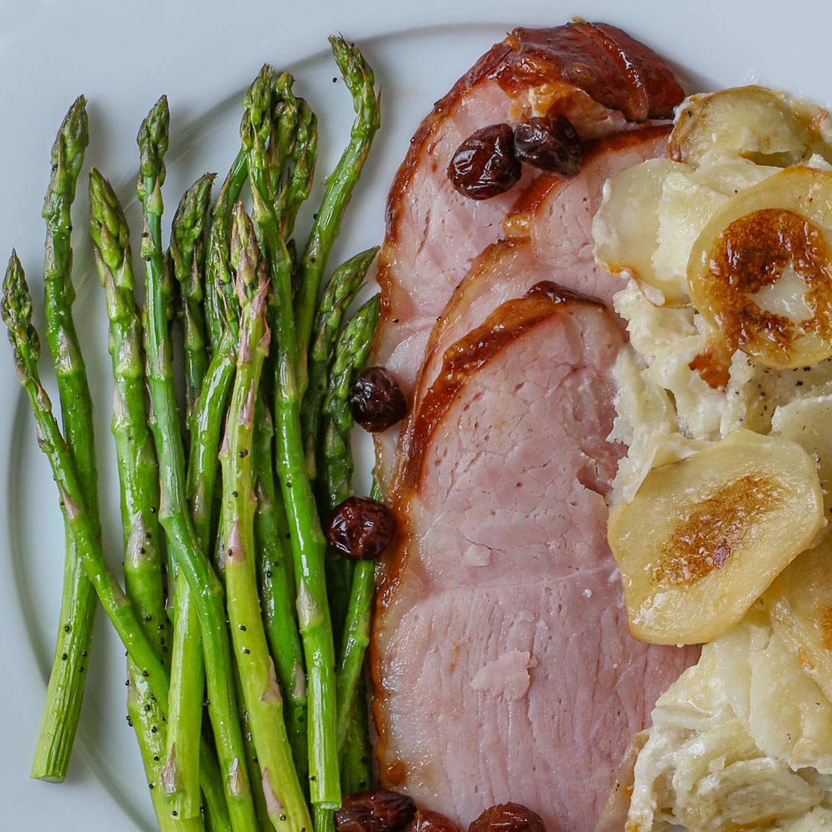 close up of a dinner plate with crock pot ham, cranberry garnish, asparagus, and scalloped potatoes.