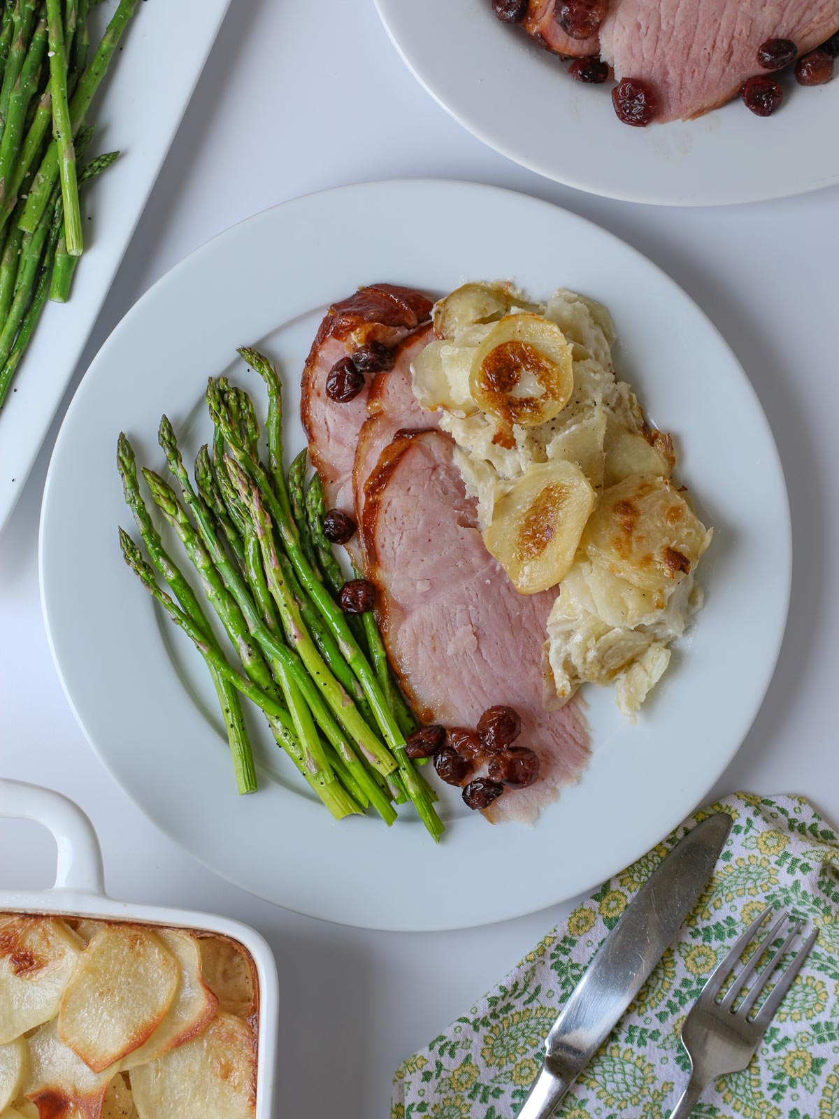 Easter dinner plate with asparagus, ham, and potatoes, with platters nearby.
