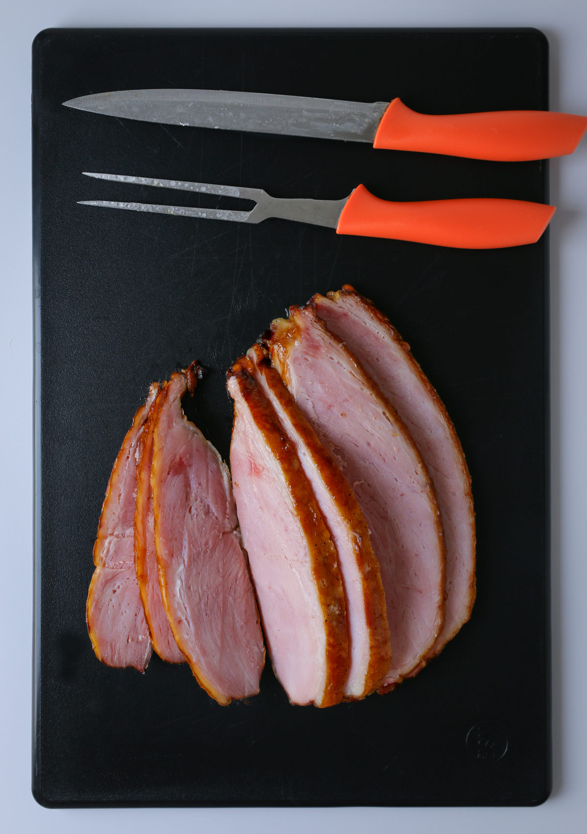 slicing ham on black cutting board with orange knife and carving fok.
