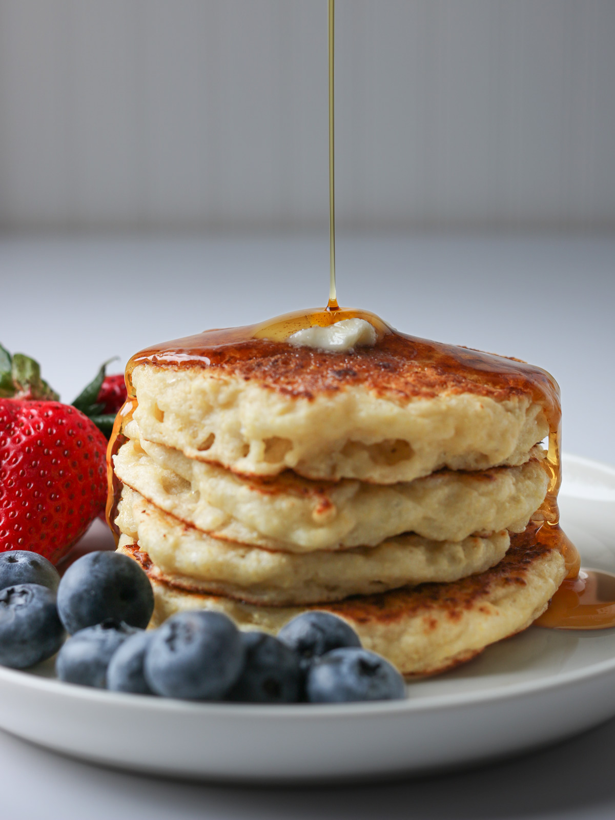 maple syrup drizzling over stack of pancakes with fresh berries on the plate.