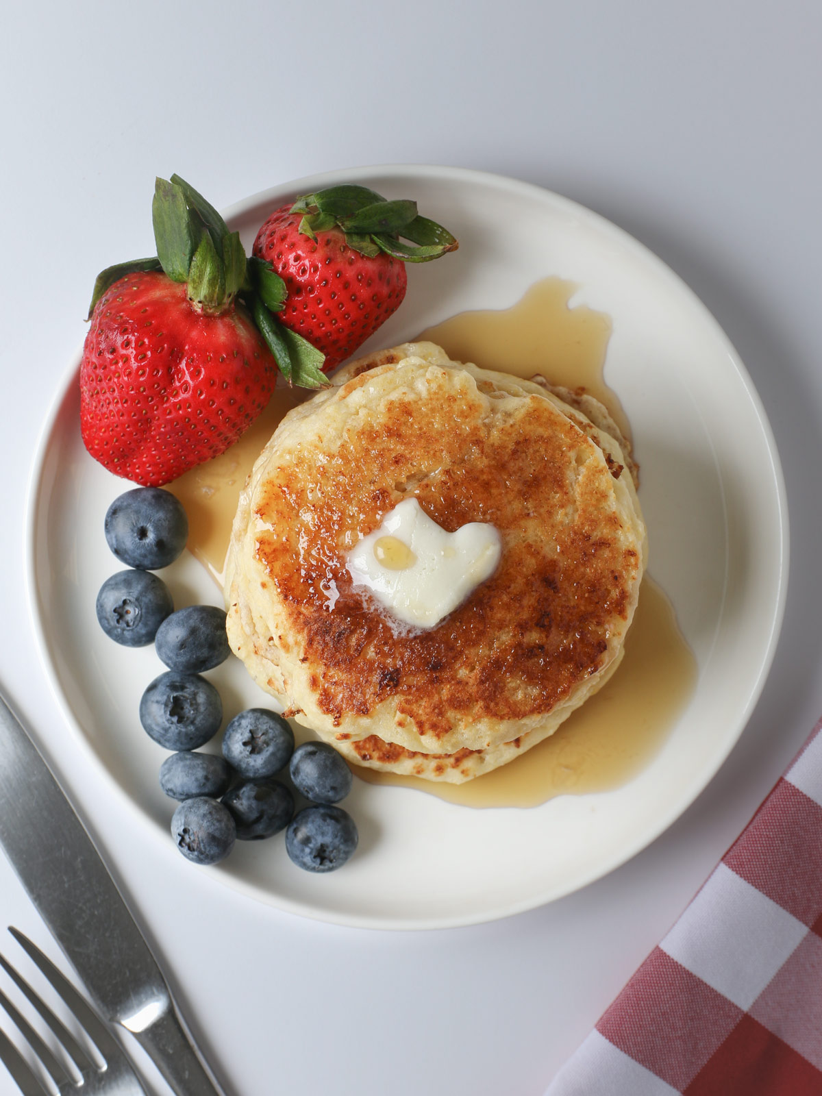 stack of pancakes on white plate with berries, near a red checked napkin with flatware.