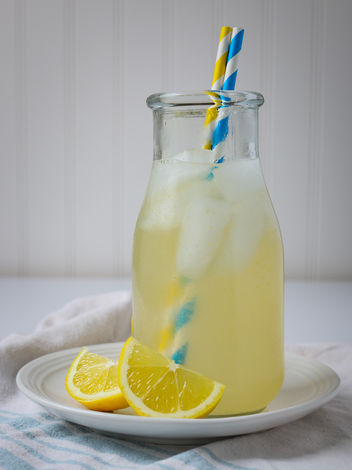glass milk jar full of ginger lemonade with blue and yellow straws, ice, and lemon wedges.