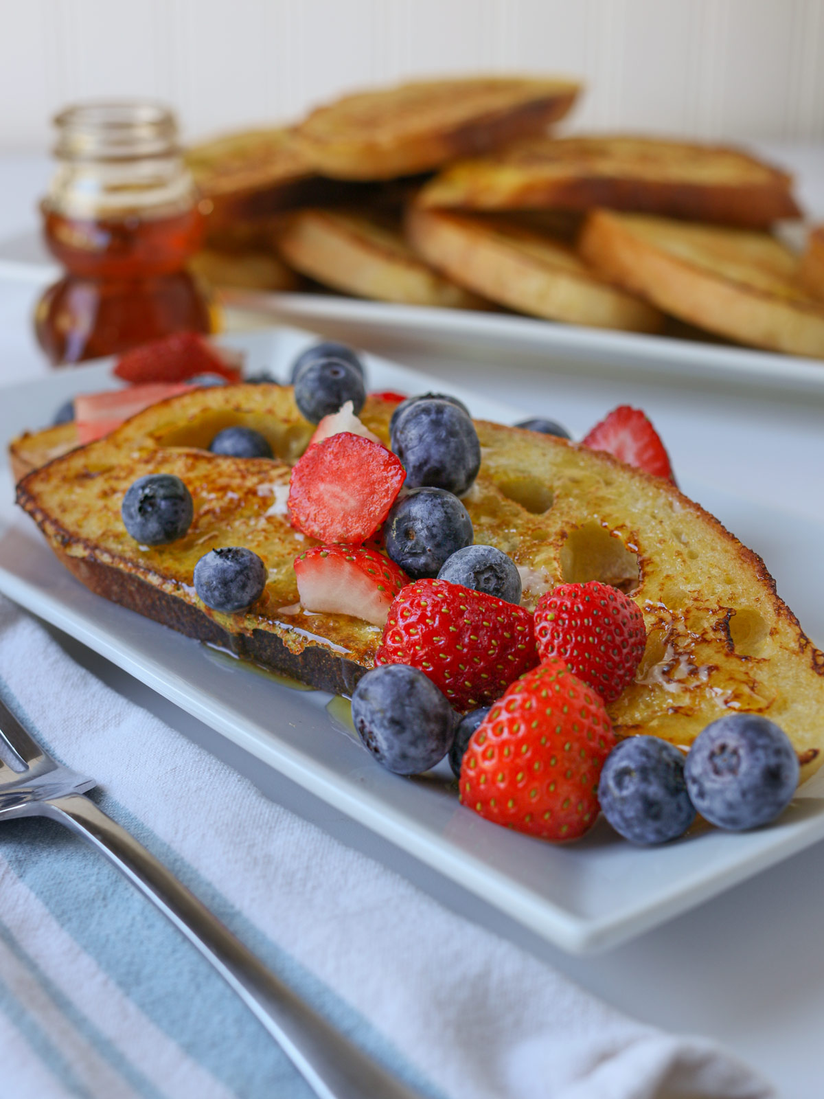 plate of sourdough french toast topped with berries near a bear jar of honey and a platter of more french toast.