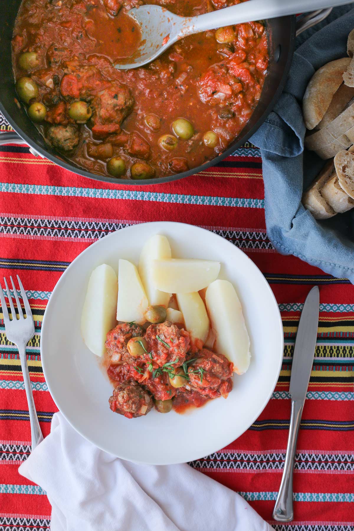 dinner table set with pot of meatballs, bread basket, and dinner plate dished up with cutlery, all atop a striped tablecloth.