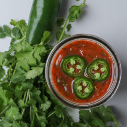 jar of jalapeño salsa with sliced chiles on top and bunch of cilantro and whole on table beside.