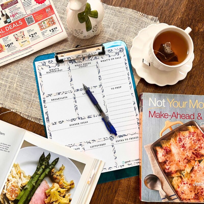array of items on table: grocery ad, cup of tea, ceramic duck, meal planning worksheet, and cookbooks.