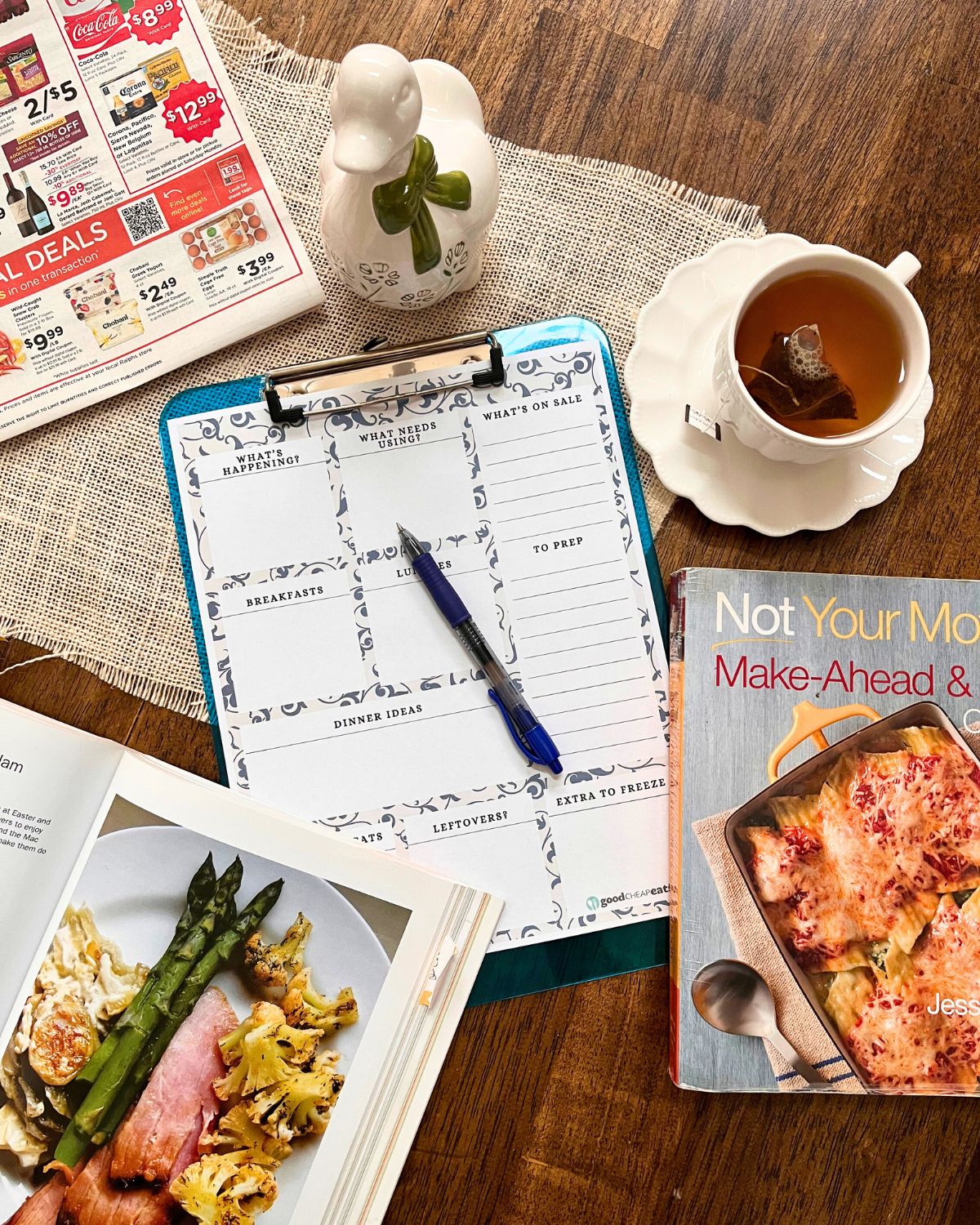 tablescape of grocery sales ad, cup of tea, clipboard with meal plan, pen, and cookbooks, with a china duck in the center.