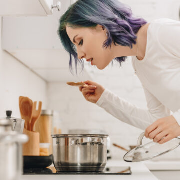 girl with purple blue hair in kitchen tasting something she's cooked in a pot on the stove.