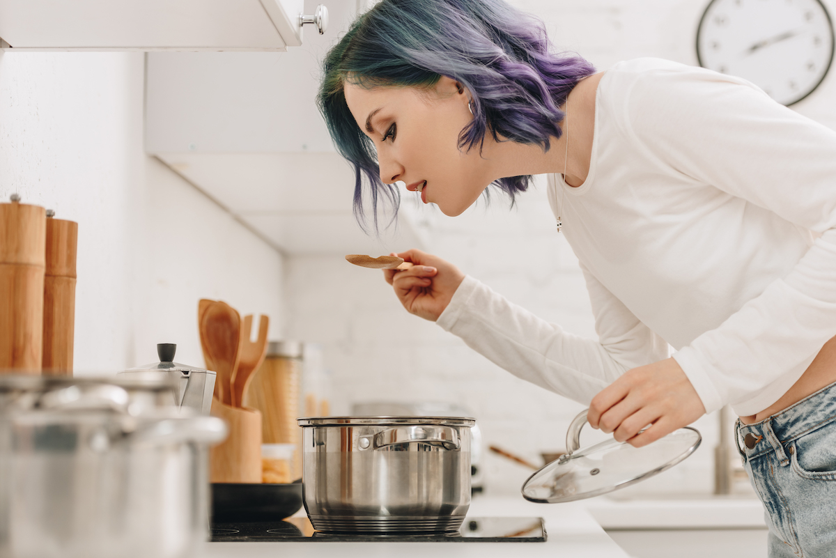 girl with purple blue hair in kitchen tasting something she's cooked in a pot on the stove.