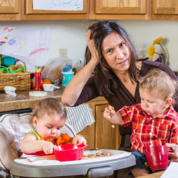 harried mom holding boy at kitchen table while girl sits in high chair nearby.