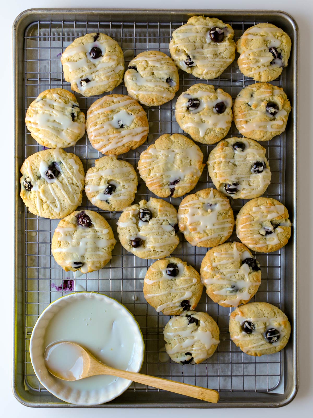 blueberry cookies on a rack set into a baking sheet with the dish of glaze and the wooden spoon nearby.