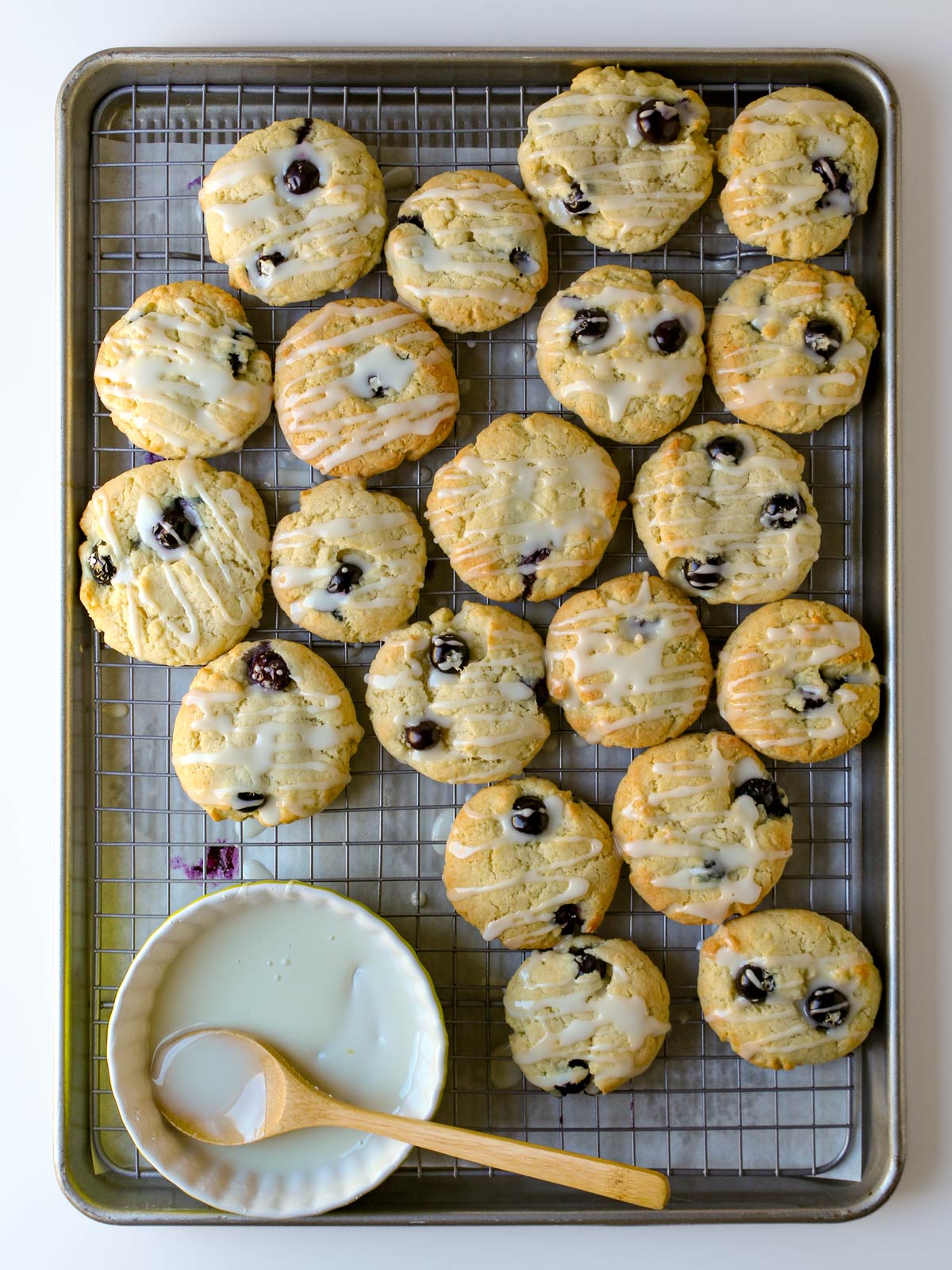 lemon blueberry cookies on cooling rack in a tray with a dish of glaze and a spoon.