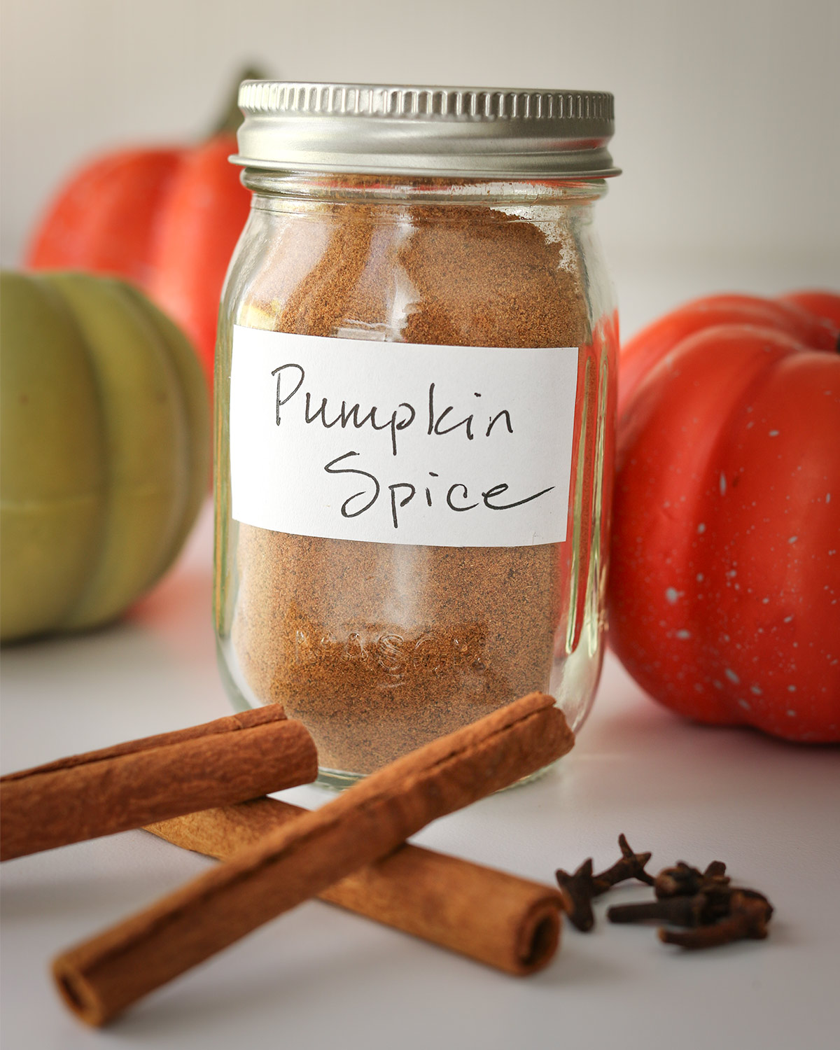 jar of pumpkin pie spice on table with small pumpkins, cinnamon sticks, and cloves.