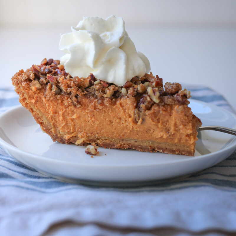 close up of slice of pumpkin pie with whipped cream on plate atop blue striped napkin.
