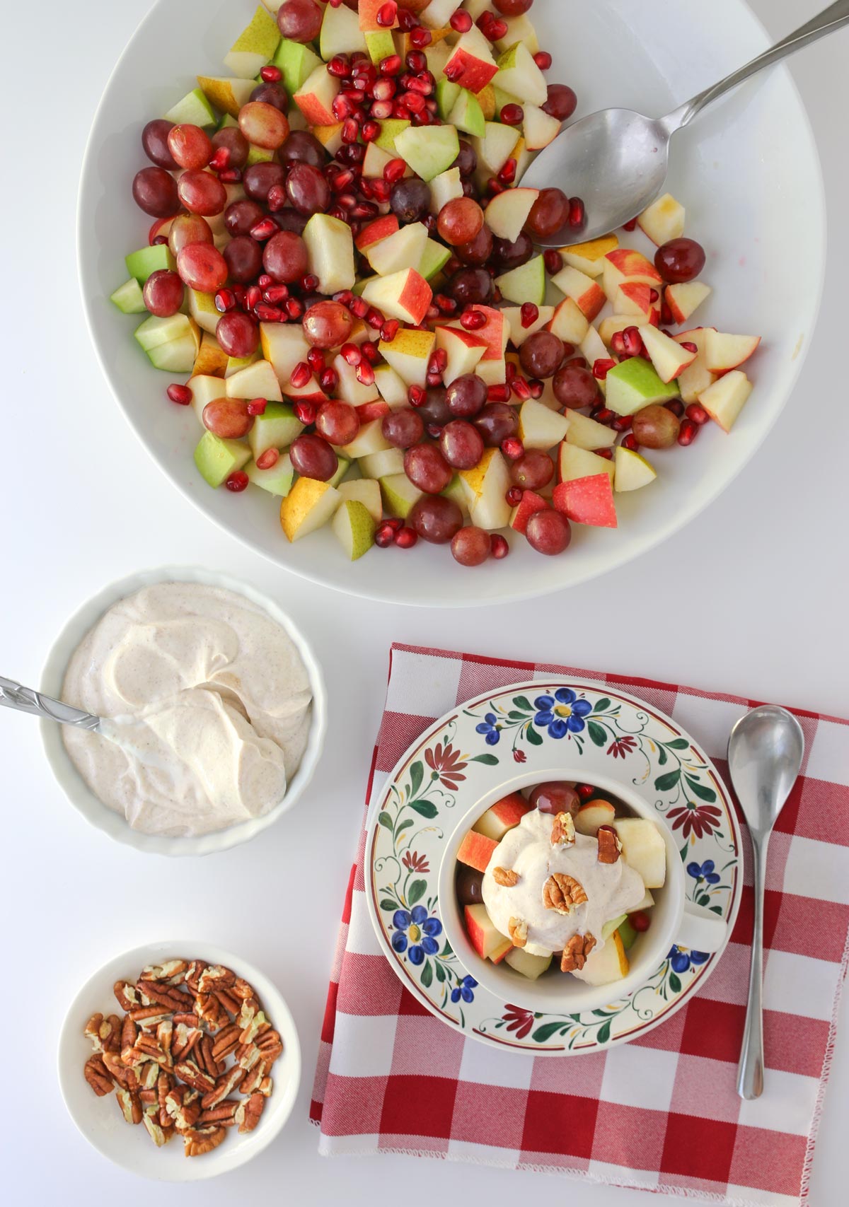 bowl of fruit with smaller bowls of yogurt topping and chopped pecans with a prepared dish on a table with a red checked cloth.