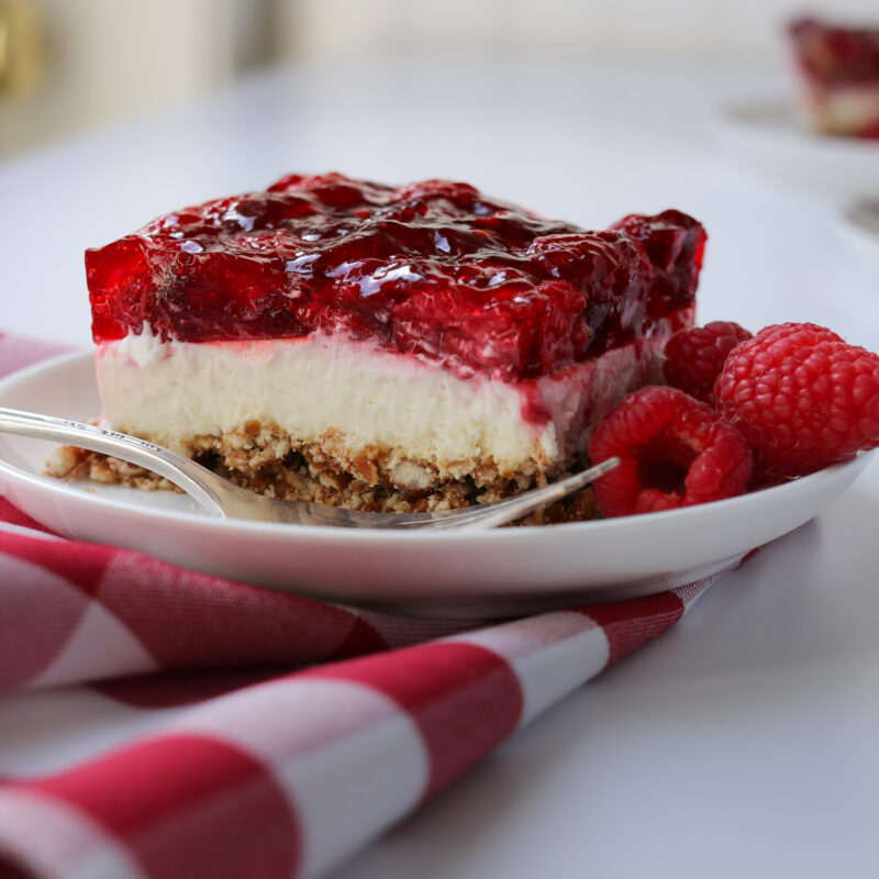 side view of a square of rapsberry pretzel dessert on a white plate with fresh berries, a fork, and a red-checked napkin.