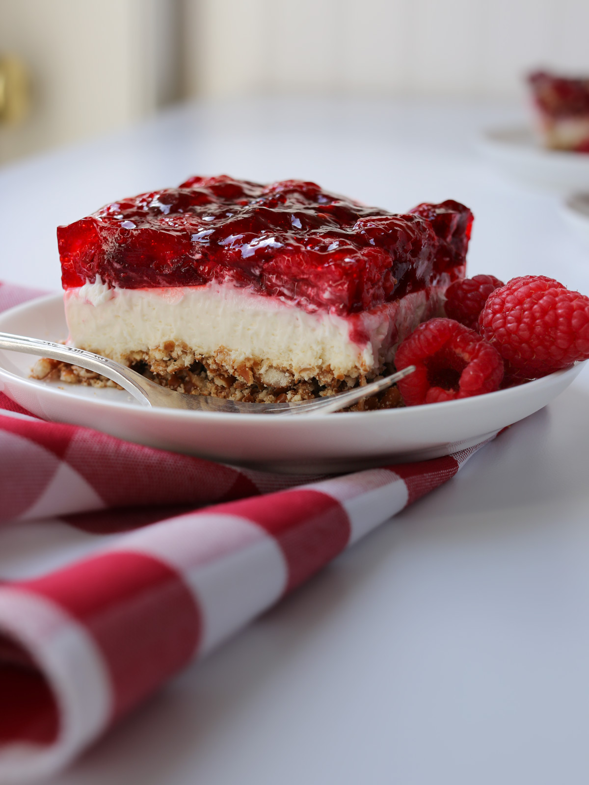 layered pretzel jello dessert on a plate with a red and white checked cloth on a white table.