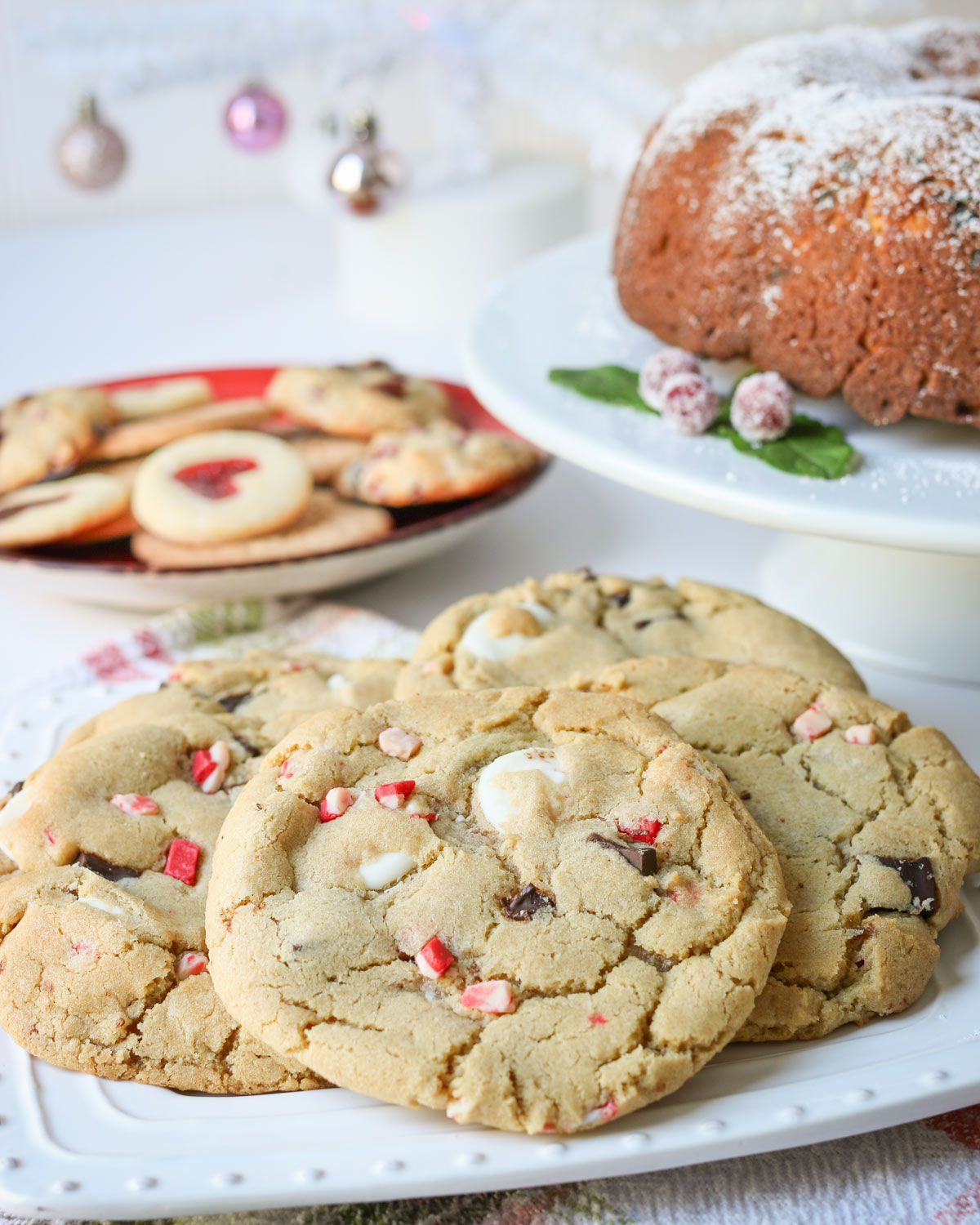 platter of peppermint chocolate chip cookies next to Christmas bundt cake on a stand and a platter of jam cookies.