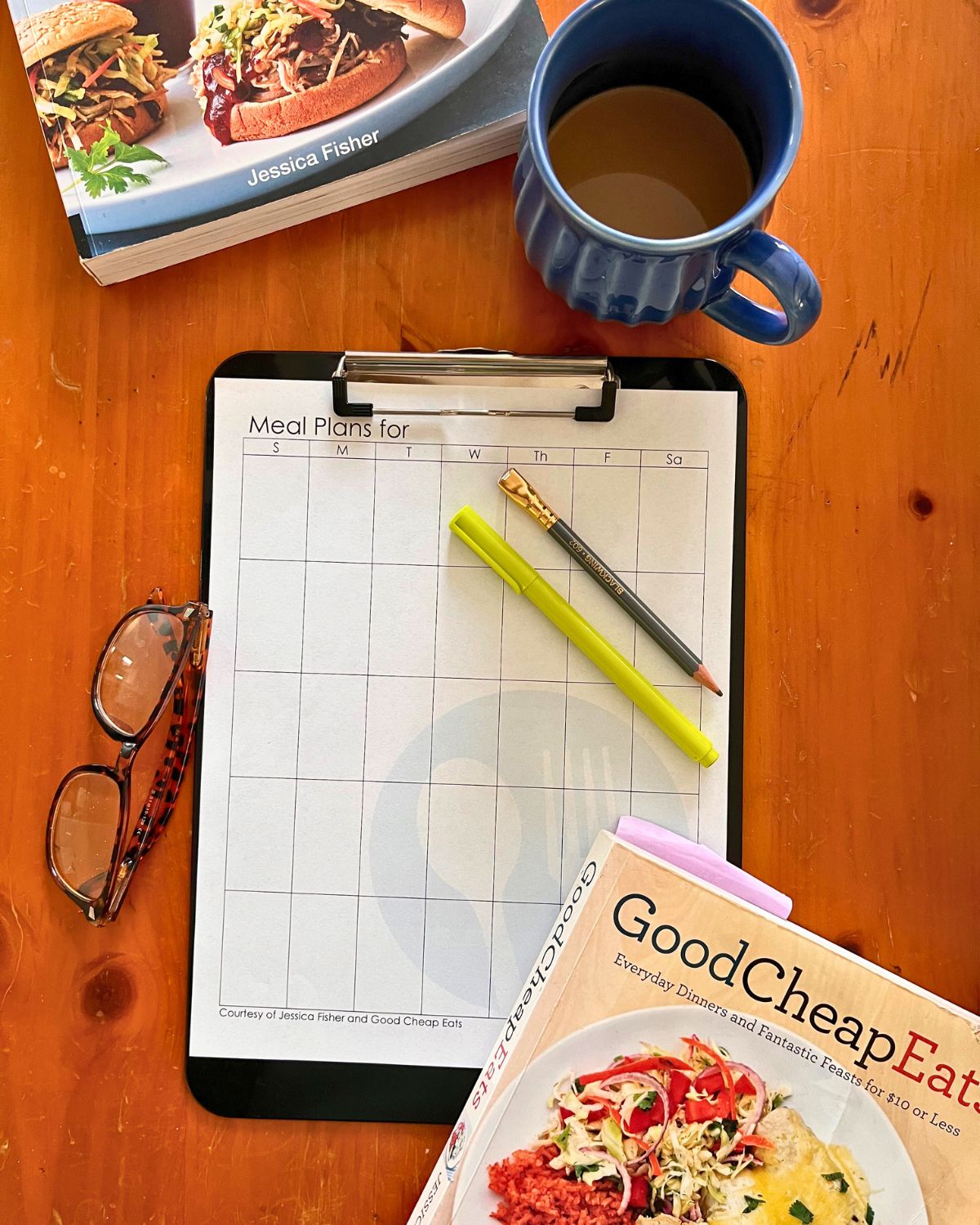 monthly meal plan printable on a clipboard with coffee, pen, pencil, glasses, and cookbooks.