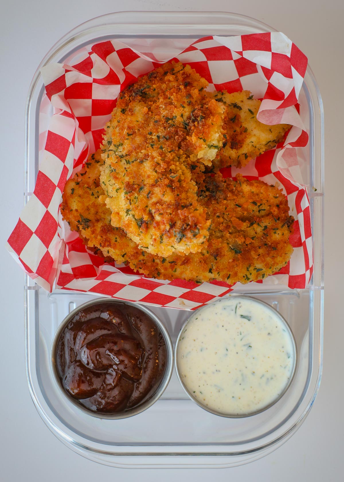 fried chicken strips in a meal prep container lined with a checked piece of parchment, with two sauce cups.