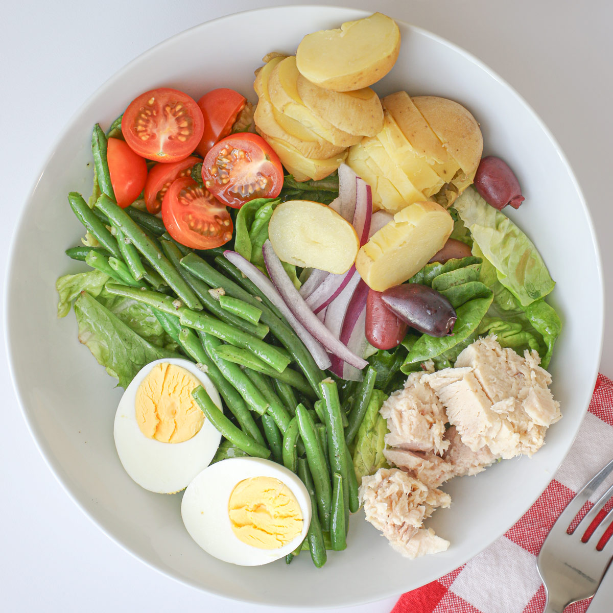 nicoise salad with green beans in a white bowl with red checked napkin.