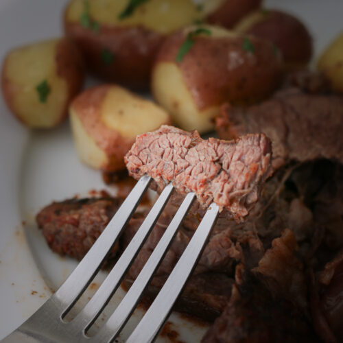 a bite of pot roast on a fork with meat and potatoes in the background.