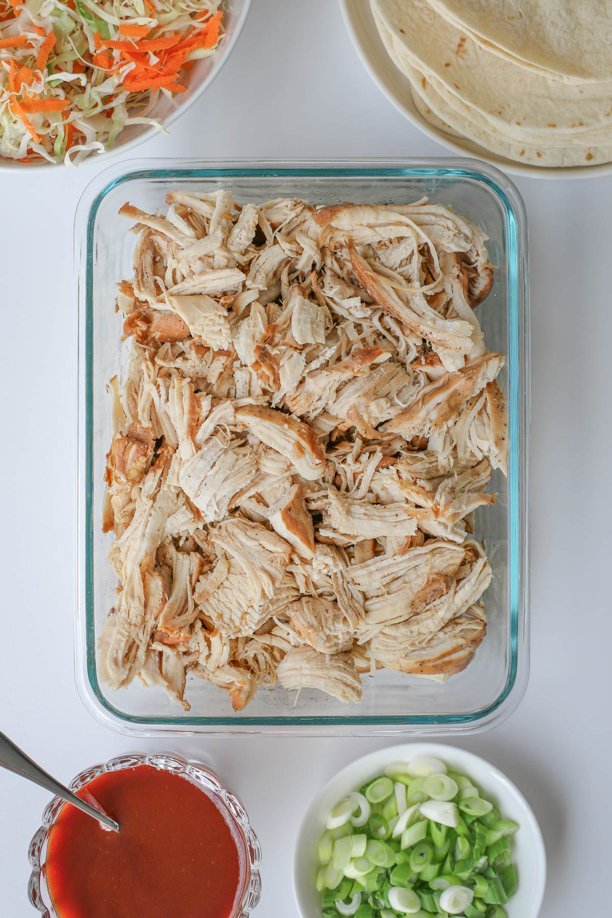 cooked shredded chicken in a glass dish with moo shu elements on dishes surrounding it.