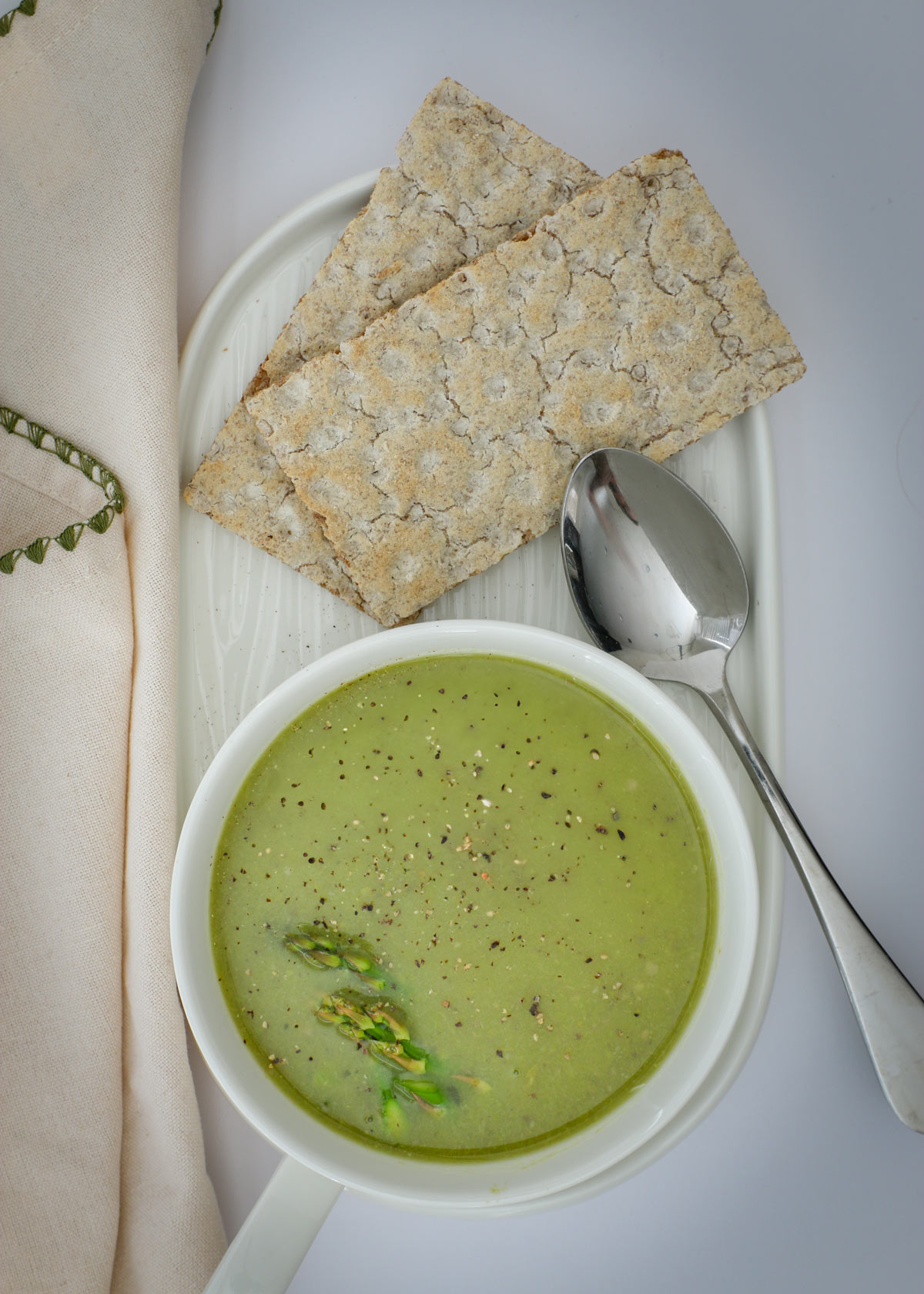 asparagus soup in a white dish with a spoon and crackers on the side with a beige napkin.