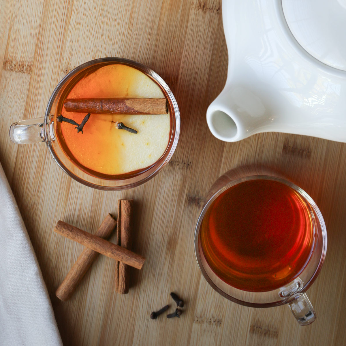 overhead shot of cups of tea with apple slice floating in one, tea pot, and cinnamon sticks nearby.