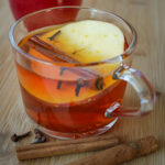 side view of glass mug filled with tea and garnished with apple slice, cinnamon stick, and cloves.