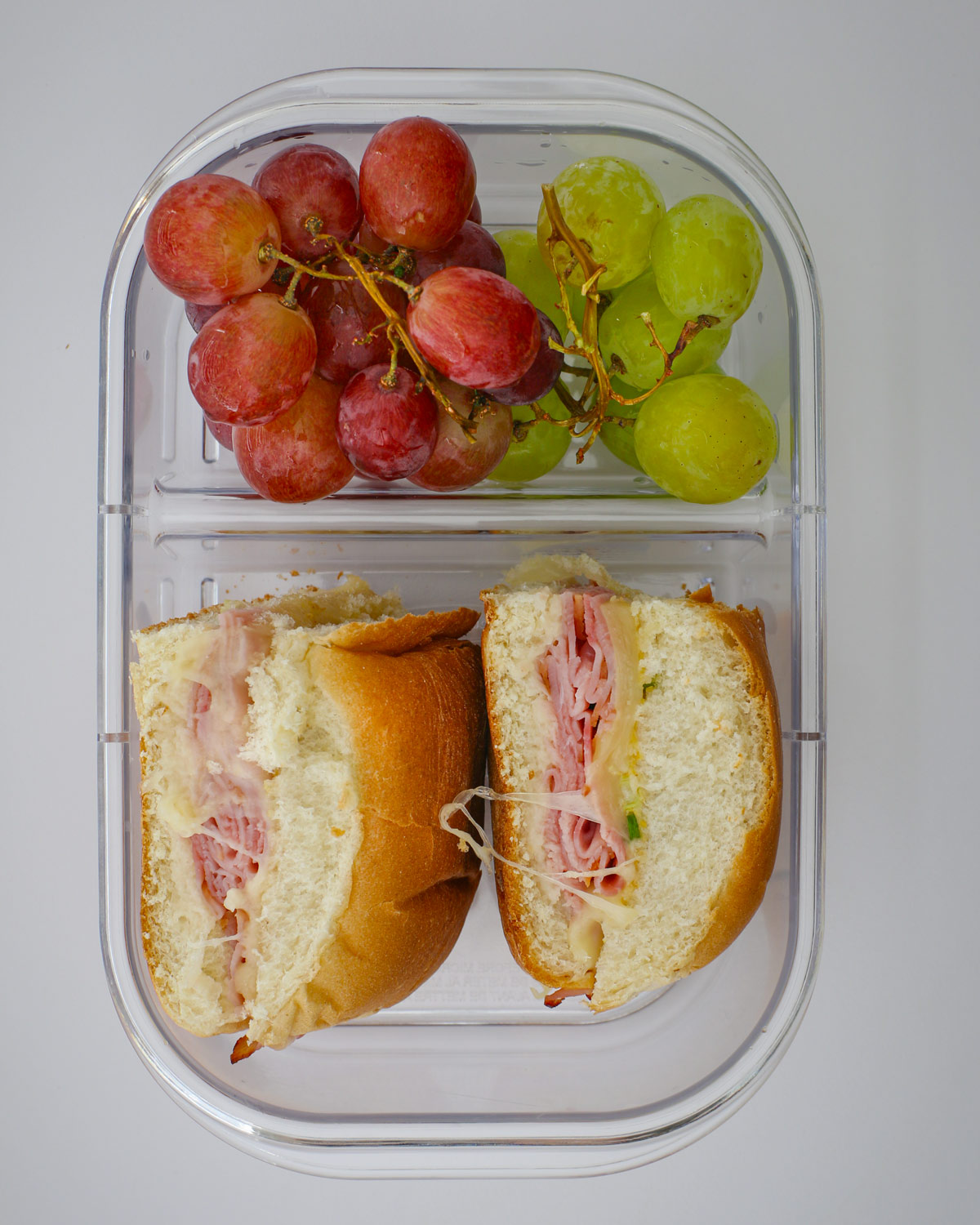 two ham and cheese sandwiches in a meal prep box with fruit.