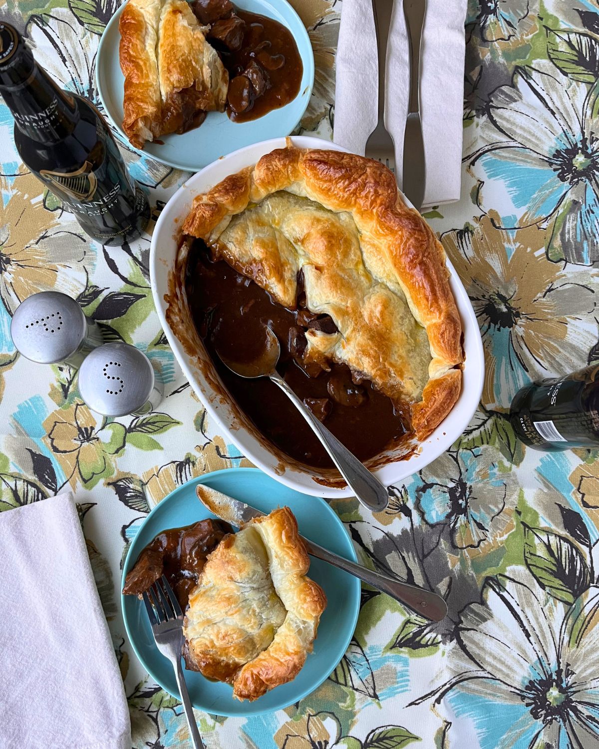 beef pot pie and two plates dished up on flowered table cloth with napkins and flatware.