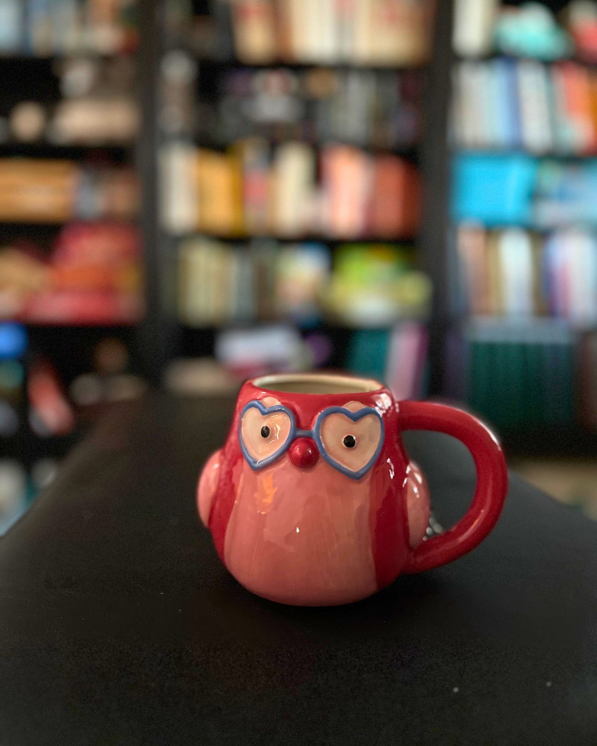 owl wearing heart glasses mug on ottoman with bookshelves in background.