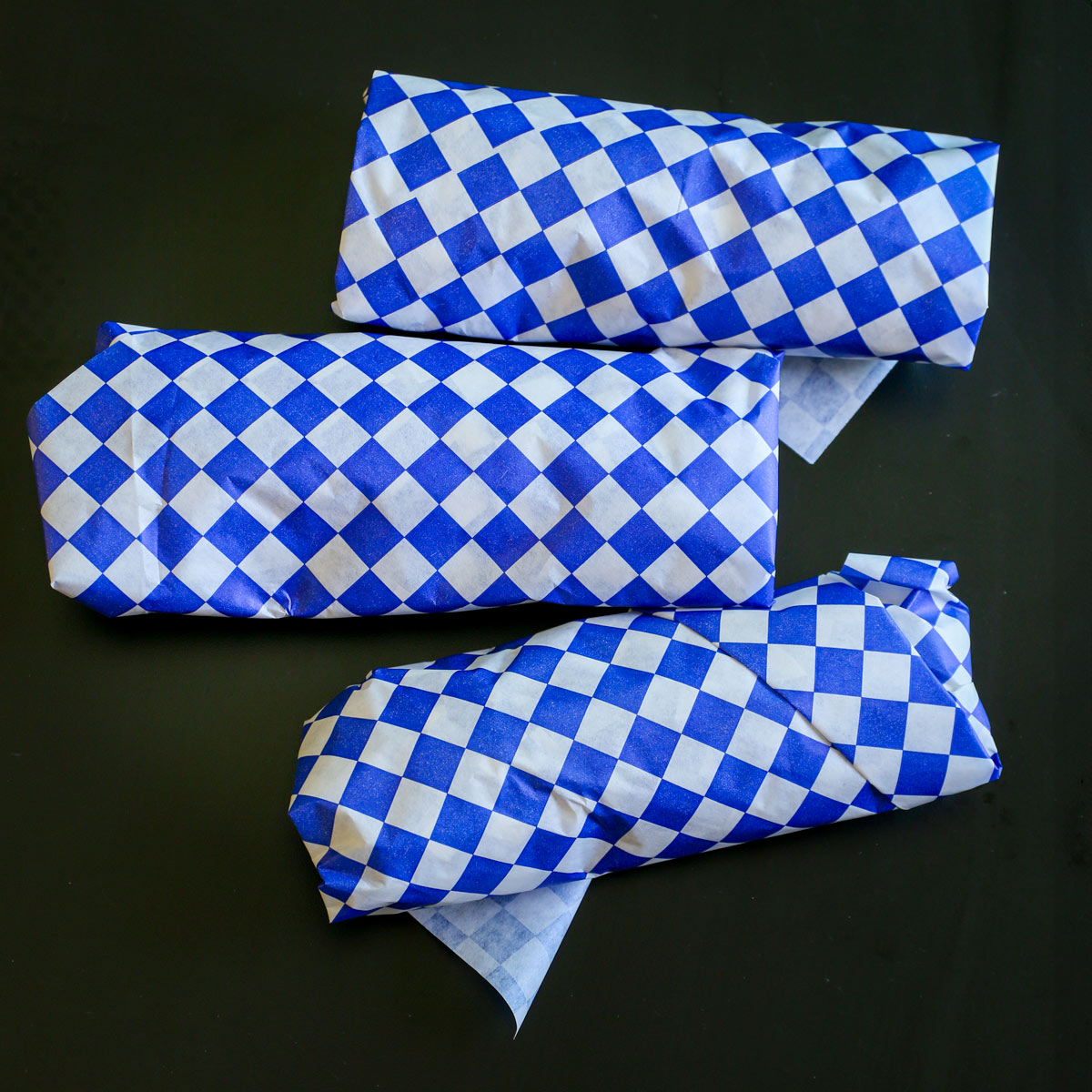 three freezer burritos wrapped in blue checked paper on black table.