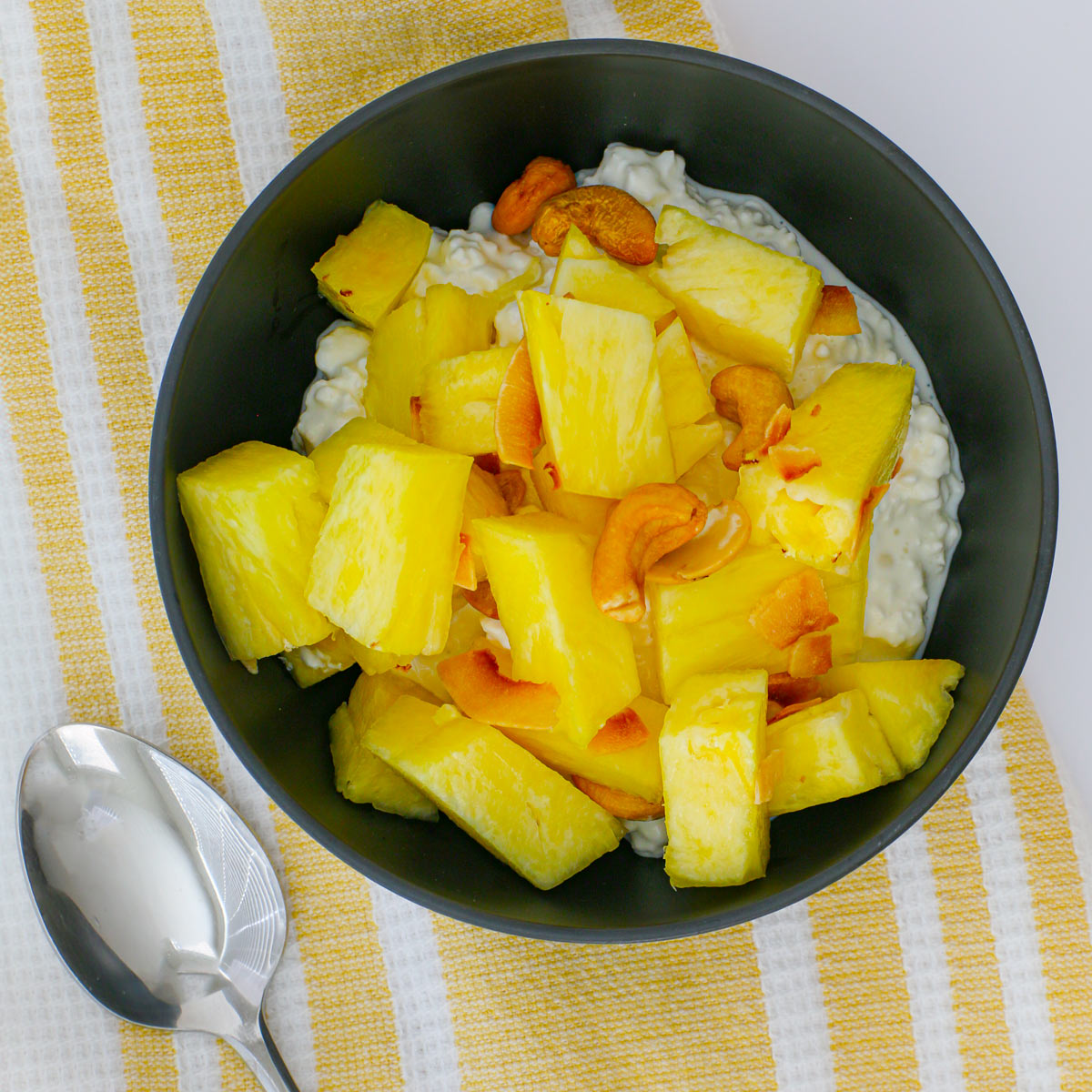 overhead shot of pineapple salad over cottage cheese in black bowl on yellow towel with spoon on the side.
