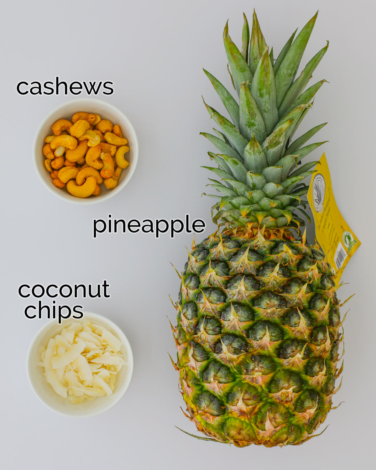 whole pineapple and bowls of cashews and coconut on a white table.