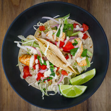 black dish with two mahi mahi fish tacos and two lime wedges.