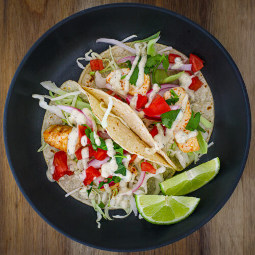 black dish with two mahi mahi fish tacos and two lime wedges.