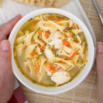 hands holding a white bowl of chicken noodle soup.