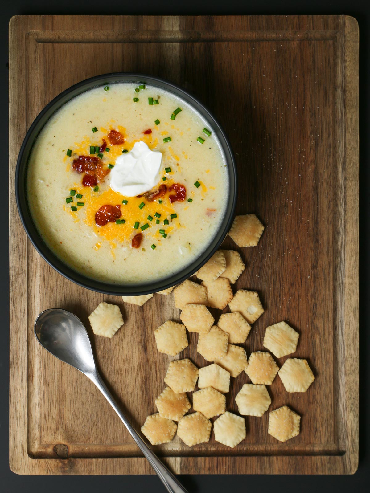 oyster crackers scattered on a wood board next to a bowl of potato soup with a spoon.