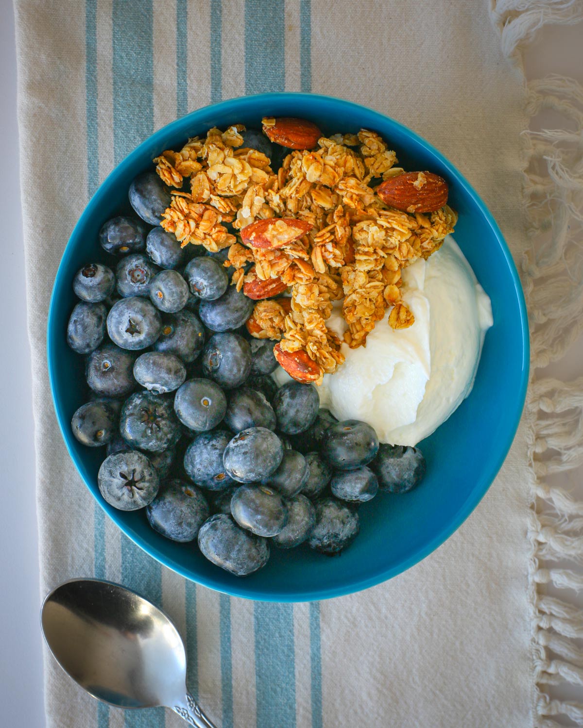 blue bowl with yogurt, berries, and vanilla almond granola with a spoon beside.