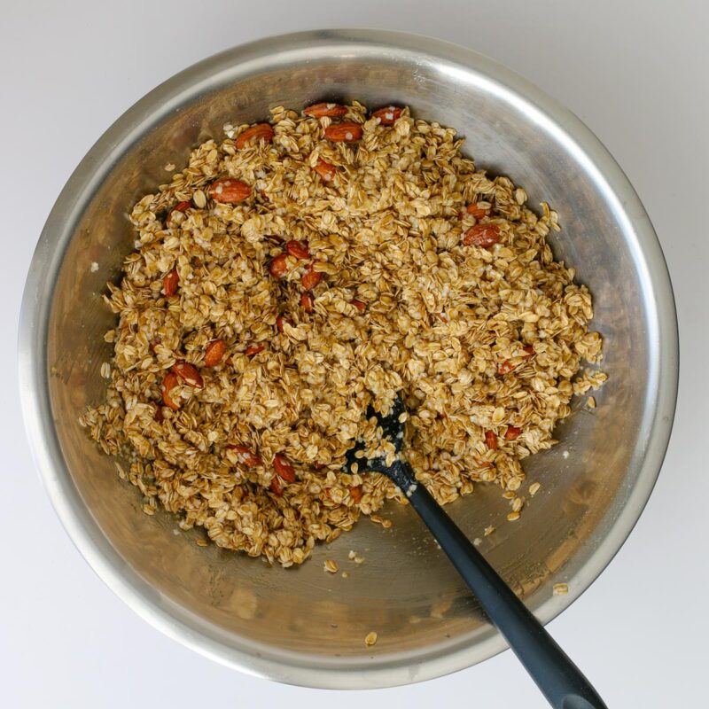 the wet granola mixture stirred together with a spatula in the stainless steel bowl.
