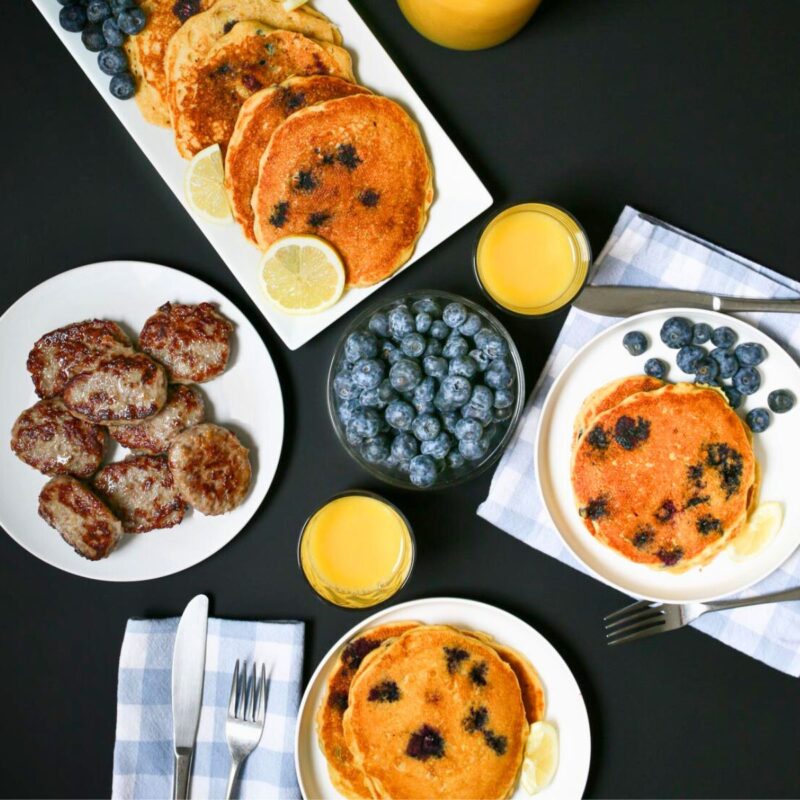 array of pancakes on platter with sausage and fruit as well as orange juice.