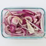 square crop of red pickled onions in glass dish with tongs.
