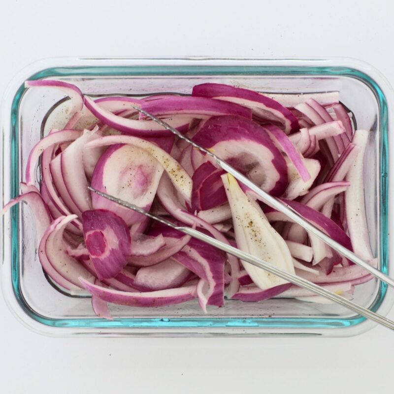 square crop of red pickled onions in glass dish with tongs.